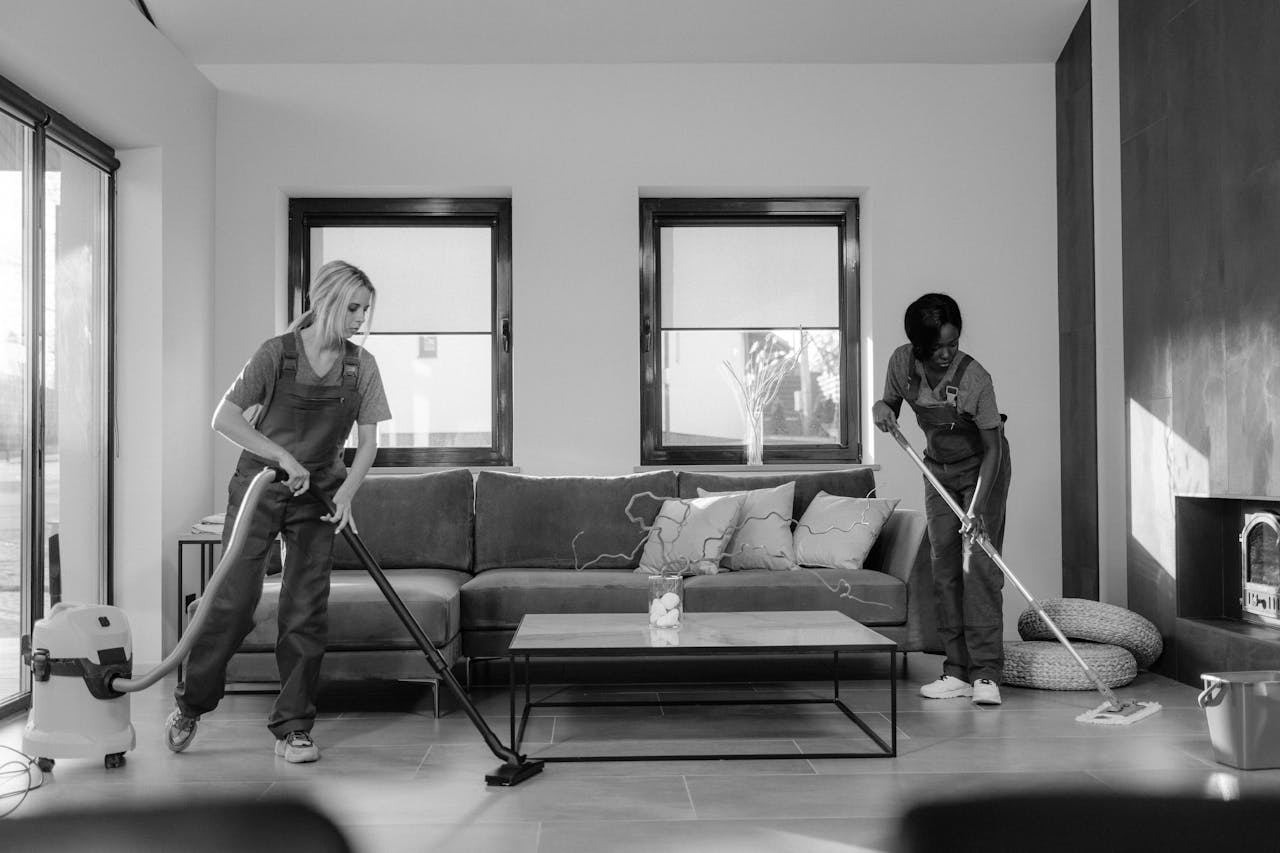 gallery-6 Two women in work clothes cleaning a modern living room using a vacuum and mop.